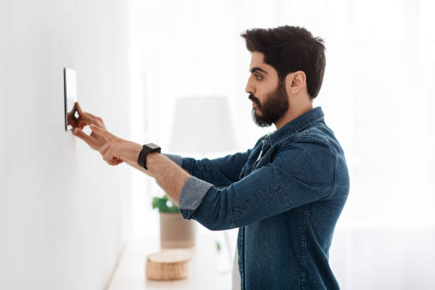 Automation domestic system. Arab man using digital tablet on wall with smart home control system app, side view Automation domestic system. Arab man using digital tablet on wall with smart home control system app, adjusting house settings, standing in light living room, side view Smart Alarms stock pictures, royalty-free photos & images